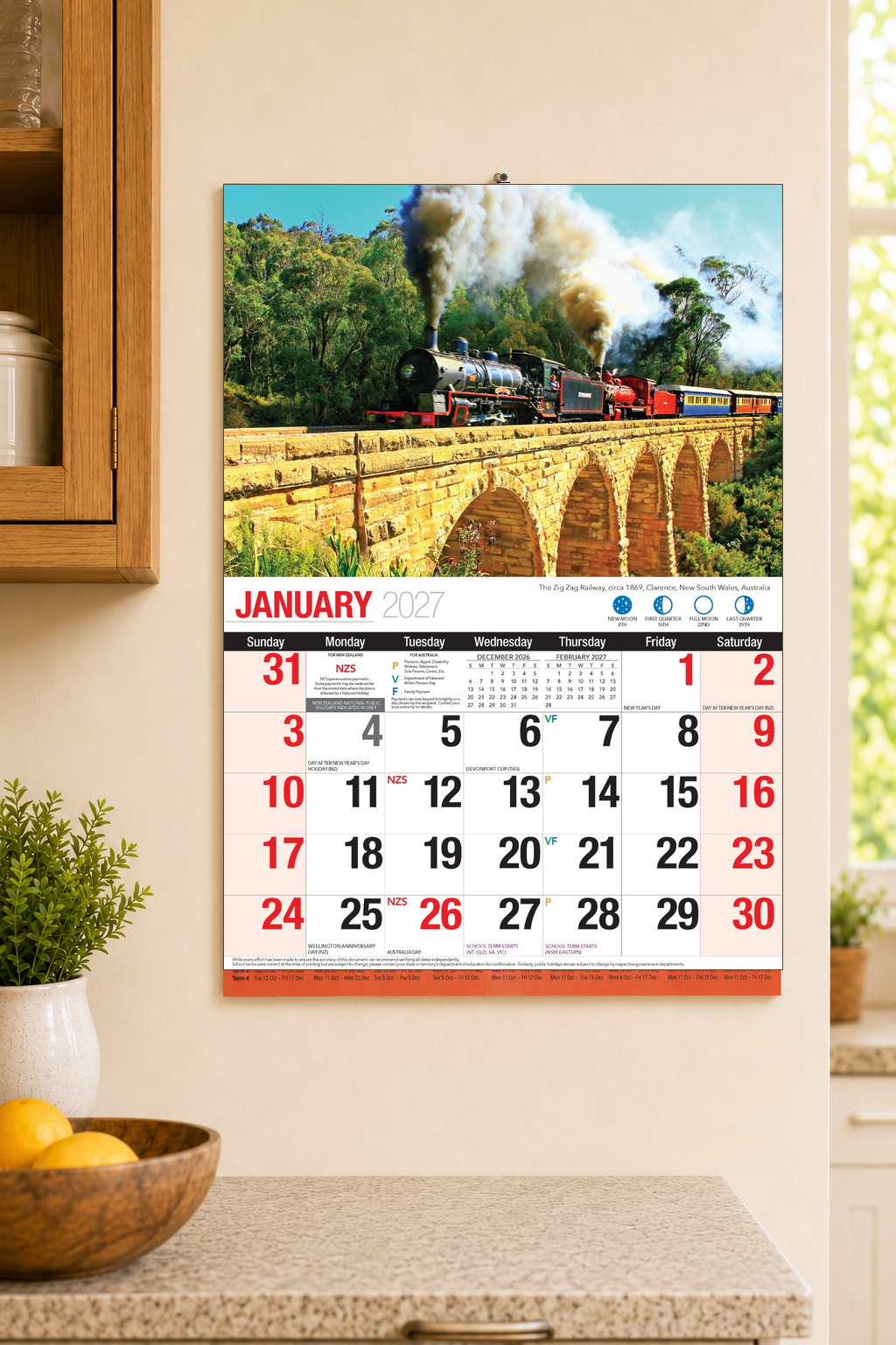 2027 Bartel Great Trains Wall Calendar displayed on kitchen wall showing January 2027 with Zig Zag Railway