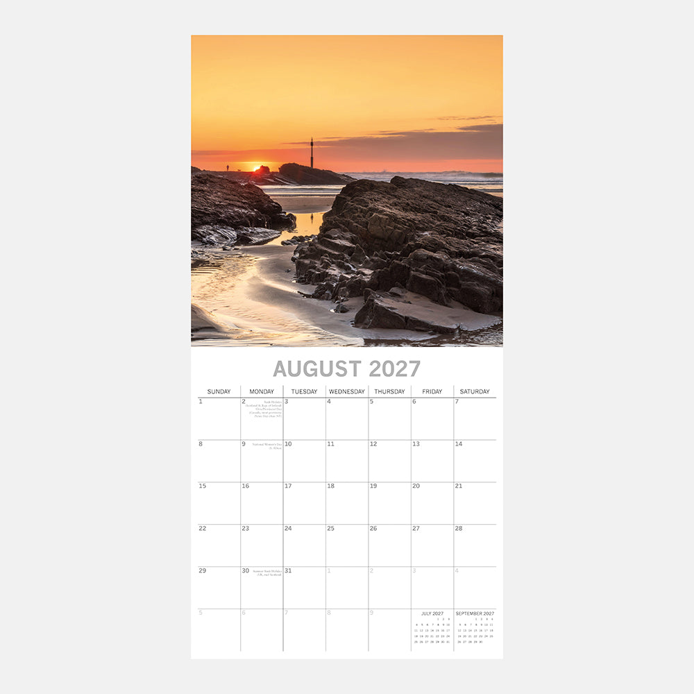 Sunsets 2027 Calendar – August 2027 page featuring golden sunset over rocky beach with lighthouse silhouette and lone figure walking on sand