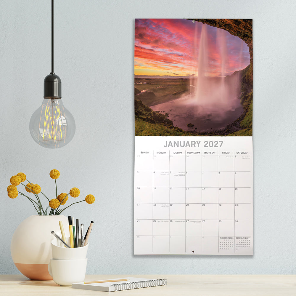 Sunsets 2027 Calendar – January 2027 lifestyle shot on grey wall above desk featuring pink and red sunset sky behind Icelandic waterfall viewed from cave
