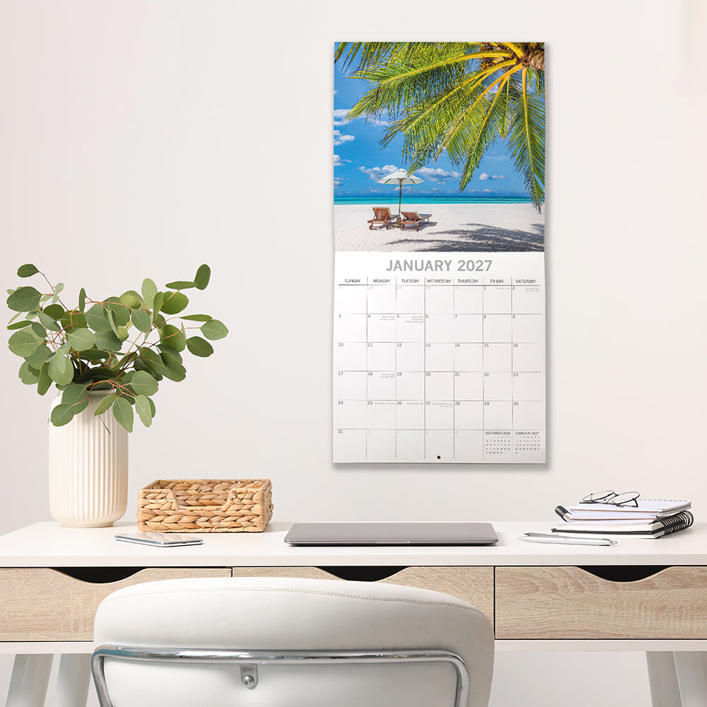 Tropical Escapes 2027 Calendar – January 2027 lifestyle shot on white wall above desk featuring two sun loungers and white umbrella on powder-white tropical beach