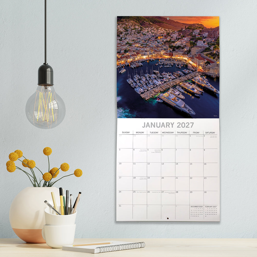 Greek Islands 2027 Calendar - lifestyle shot on grey wall above desk showing January 2027 page with aerial night view of Hydra marina packed with superyachts lit up at sunset with orange sky