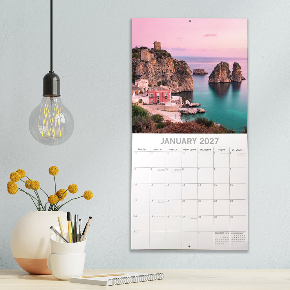 Mediterranean 2027 Square Wall Calendar - January 2027 lifestyle shot hung on wall above a styled desk with Scopello Sicily bay at sunset