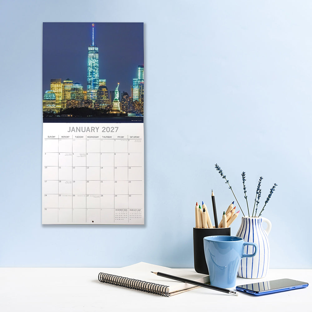 New York 2027 Calendar – January 2027 lifestyle shot on blue wall above desk featuring Statue of Liberty and One World Trade Center illuminated against Lower Manhattan skyline at night