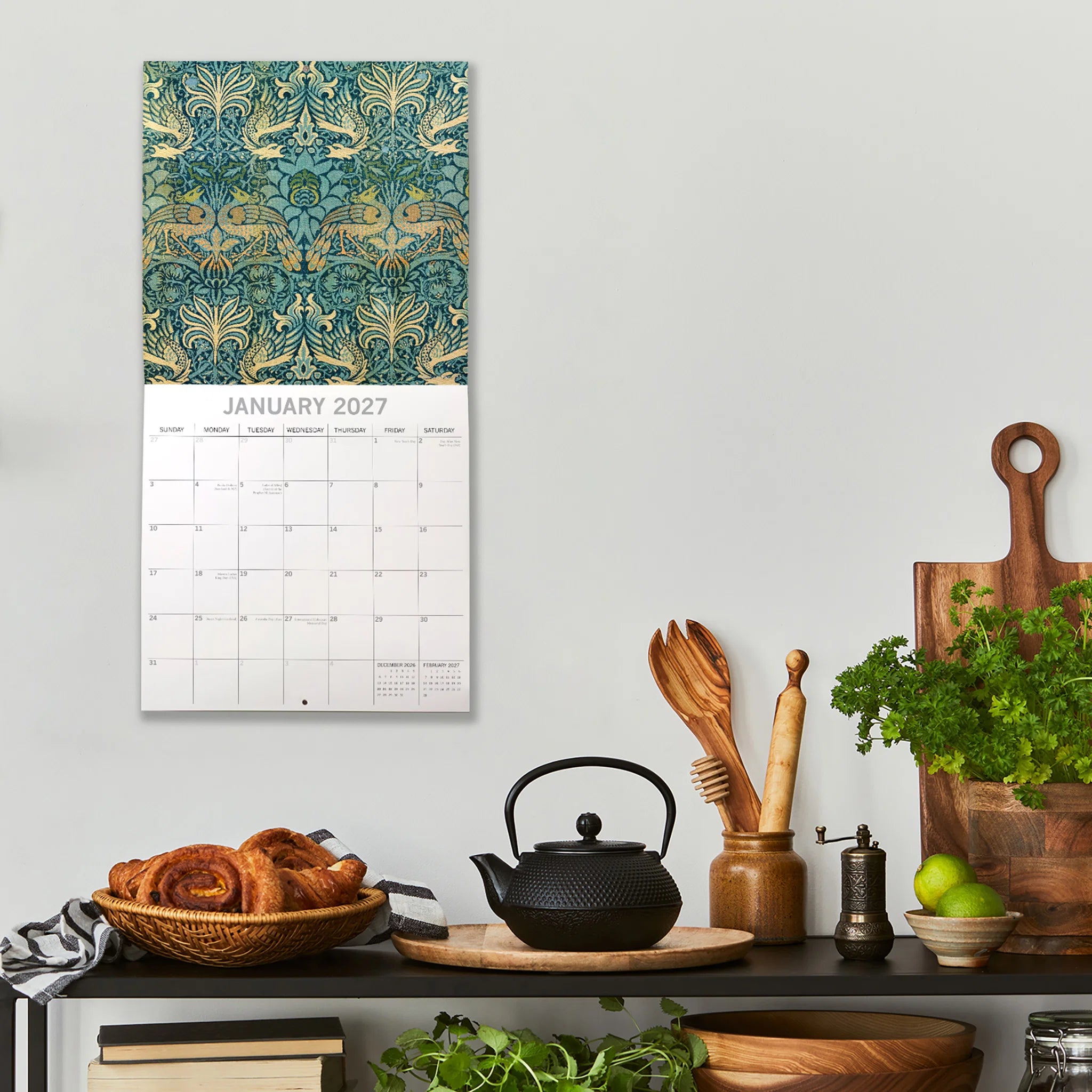 William Morris 2027 Calendar - lifestyle shot on kitchen wall showing January 2027 page with Peacock and Dragon pattern in teal and gold above date grid