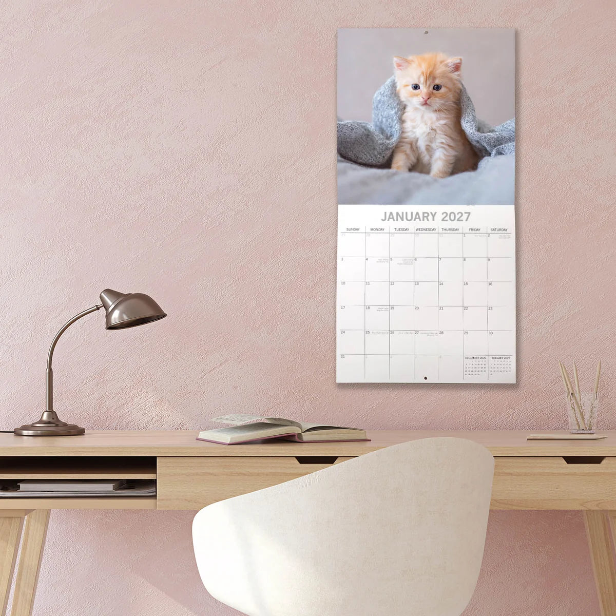 Cute Kittens 2027 Calendar - lifestyle shot on wall in pink room showing January 2027 page with cream kitten wrapped in blue blanket on grey bed