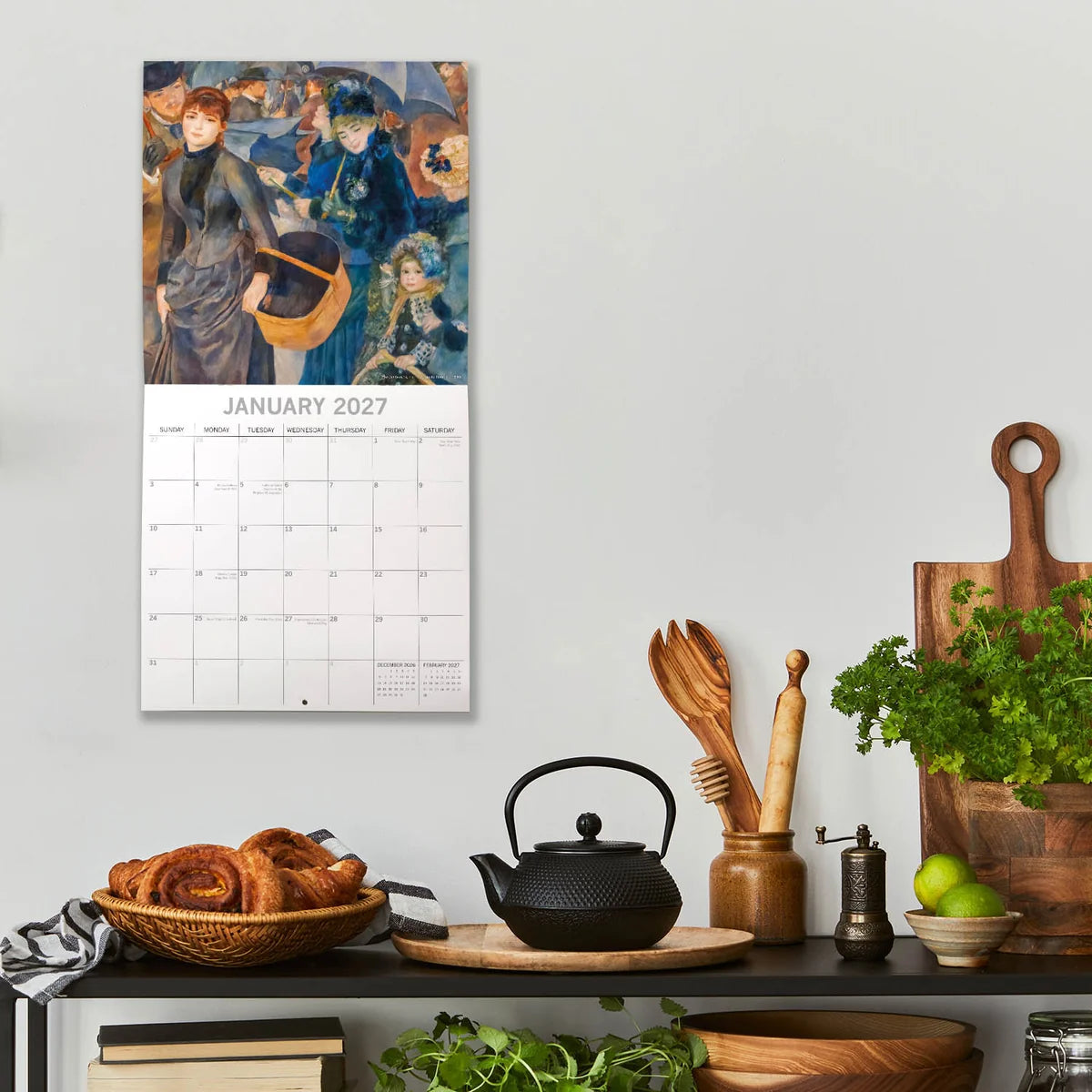 Impressionists 2027 Calendar - lifestyle shot on kitchen wall showing January 2027 page with Renoir's The Umbrellas featuring elegantly dressed Parisians with blue umbrellas in rain