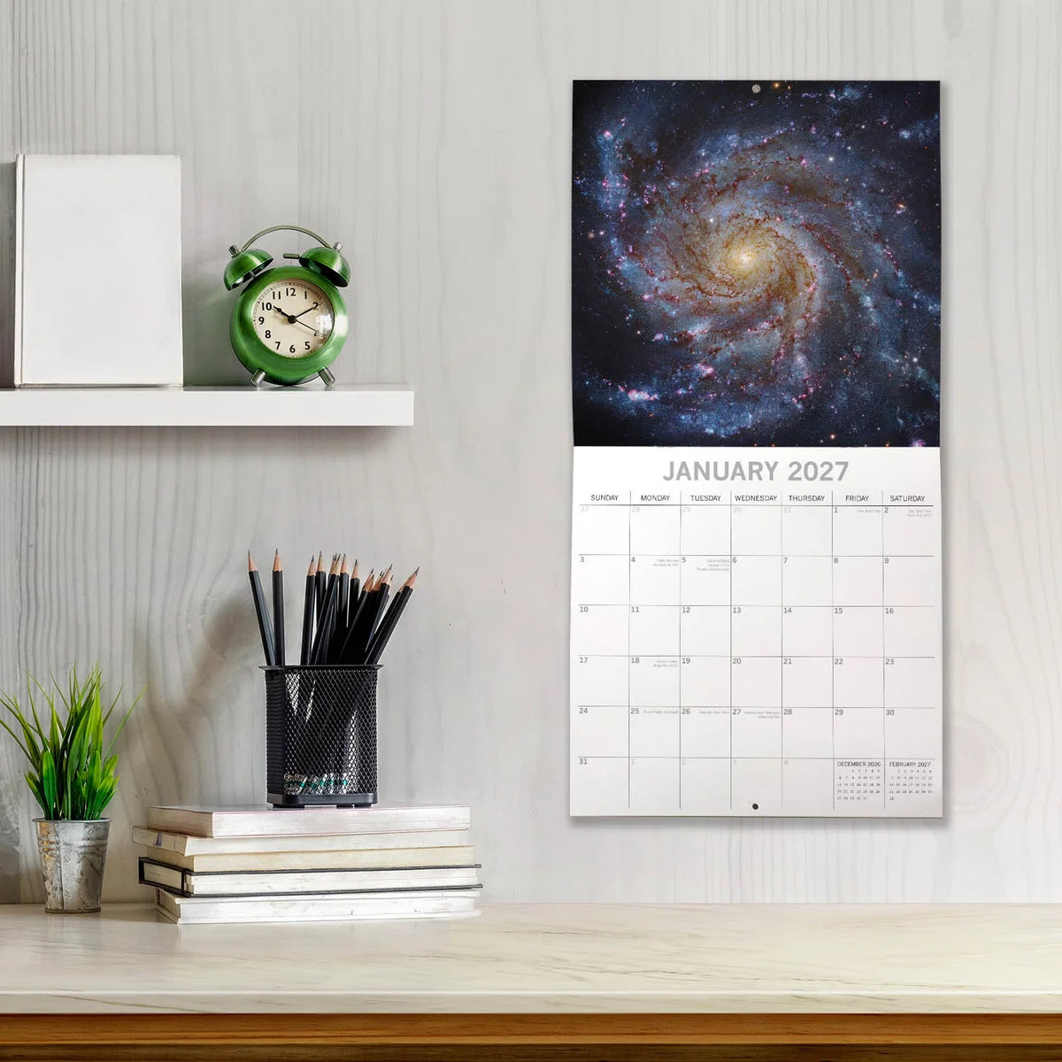 Space 2027 Calendar - lifestyle shot on white wall above desk showing January 2027 page with the Pinwheel Galaxy spiral arms in blue and gold on black starfield background