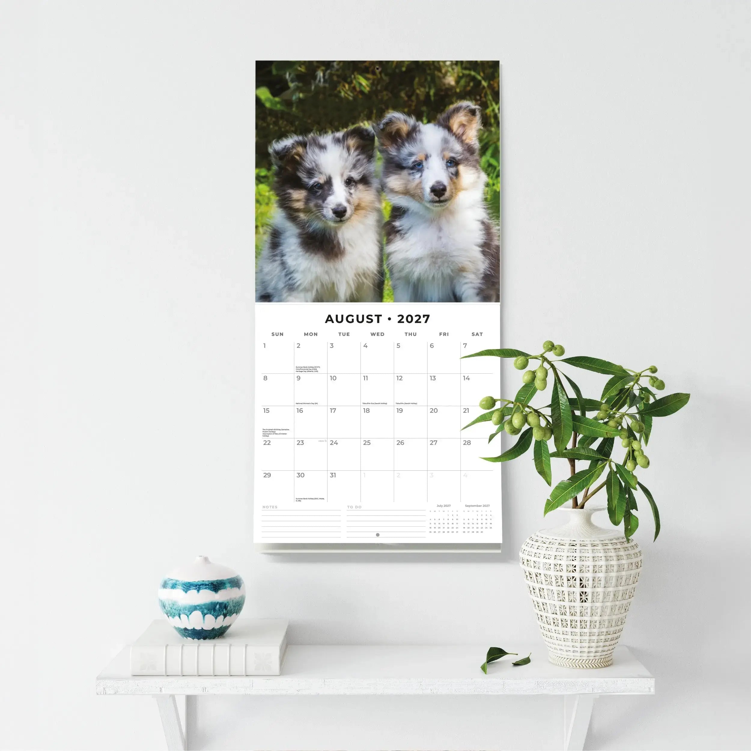 Adorable Dogs 2027 Square Wall Calendar – flat lay lifestyle shot showing front cover with two puppies playing in grass alongside coffee notebook and pen on marble surface